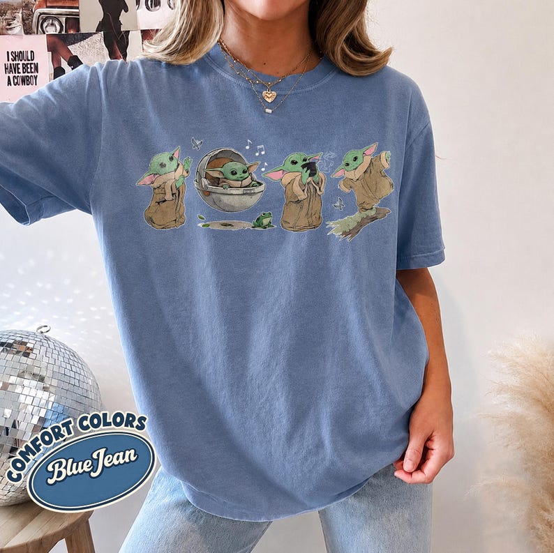 May include: A blue jean colored t-shirt featuring a graphic print of a small green creature in various poses, including sitting in a floating pod, holding a cup, and standing on a log, accompanied by a small green frog and musical notes.