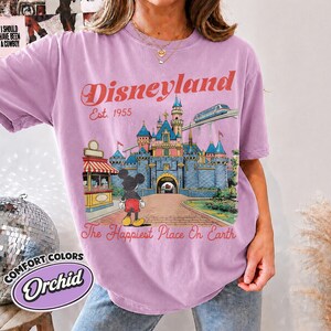 May include: An orchid-colored t-shirt featuring a graphic of the Disneyland castle, Mickey Mouse, and a monorail. The text reads Disneyland Est. 1955 and The Happiest Place On Earth. The design includes a red and white striped kiosk and blue castle.