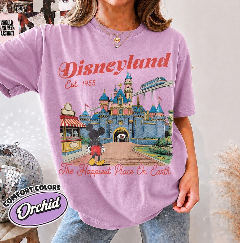 May include: An orchid-colored t-shirt featuring a graphic of the Disneyland castle, Mickey Mouse, and a monorail. The text reads Disneyland Est. 1955 and The Happiest Place On Earth. The design includes a red and white striped kiosk and blue castle.