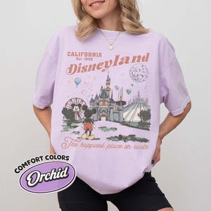 May include: Orchid-colored t-shirt with a Disneyland graphic. The design features a castle, Mickey Mouse, and the text "California Disneyland Est. 1955" and "The happiest place on earth."