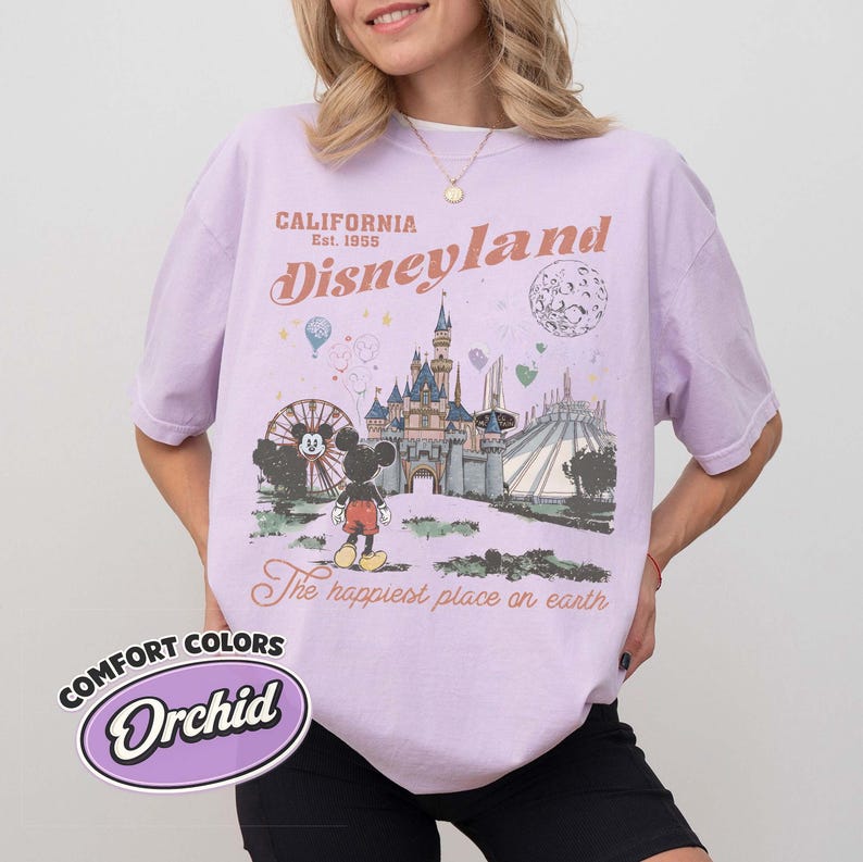 May include: Orchid-colored t-shirt with a Disneyland graphic. The design features a castle, Mickey Mouse, and the text "California Disneyland Est. 1955" and "The happiest place on earth."