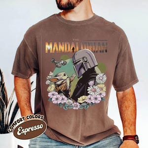 May include: Espresso-colored t-shirt featuring a graphic of The Mandalorian and Grogu surrounded by flowers. The design includes a spaceship and the text "THE MANDALORIAN". The shirt is part of the Comfort Colors collection.