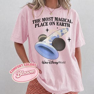 K&ouml;nnte beinhalten: Hellrosa T-Shirt mit dem Text "THE MOST MAGICAL PLACE ON EARTH" und einer Grafik eines blau-wei&szlig;en Zauberhuts mit schwarzen Mickey Mouse Ohren. Das Shirt hat auch den Text "Walt Disney World".