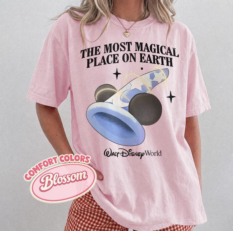 K&ouml;nnte beinhalten: Hellrosa T-Shirt mit dem Text "THE MOST MAGICAL PLACE ON EARTH" und einer Grafik eines blau-wei&szlig;en Zauberhuts mit schwarzen Mickey Mouse Ohren. Das Shirt hat auch den Text "Walt Disney World".