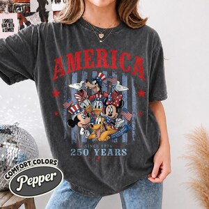 May include: A charcoal grey short-sleeve t-shirt featuring a graphic print of cartoon characters wearing patriotic hats. The text reads AMERICA, SINCE 1776, 250 YEARS, and AMERICA 250TH USA ANNIVERSARY 1776-2026. The design includes red stars and American flags.