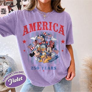 May include: A violet short-sleeve t-shirt featuring a graphic of Disney characters wearing patriotic hats. The text reads AMERICA, SINCE 1776, 250 YEARS, and USA ANNIVERSARY. The design includes red, white, and blue stars and American flags.