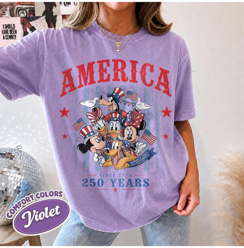 May include: A violet short-sleeve t-shirt featuring a graphic of Disney characters wearing patriotic hats. The text reads AMERICA, SINCE 1776, 250 YEARS, and USA ANNIVERSARY. The design includes red, white, and blue stars and American flags.