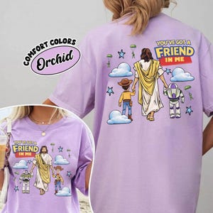 May include: Lilac Comfort Colors t-shirt with a graphic of Jesus, a child, and Buzz Lightyear, with the text "YOU'VE GOT A FRIEND IN ME." The design includes clouds, stars, and parachutes.