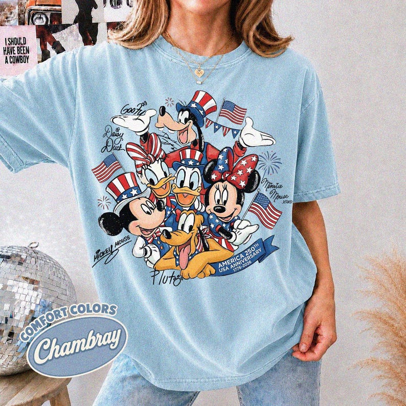 May include: Light blue chambray t-shirt featuring a graphic of Disney characters in patriotic attire, including Mickey and Minnie Mouse, Donald Duck, and Pluto. The design includes American flags and text that reads "America 250th USA Anniversary 1776-2026".