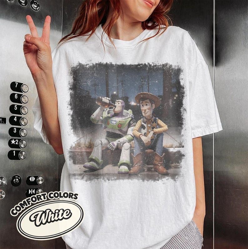 May include: White Comfort Colors t-shirt featuring a distressed graphic of Buzz Lightyear and Woody from Toy Story. Buzz is drinking from a bottle, and Woody is sitting next to him. The shirt has a crew neck and short sleeves.
