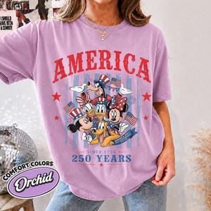 May include: An orchid-colored short-sleeve t-shirt featuring a graphic print of cartoon characters wearing patriotic hats. The text reads AMERICA, SINCE 1776, 250 YEARS, and AMERICA 250TH USA ANNIVERSARY. The design includes American flags and red stars.