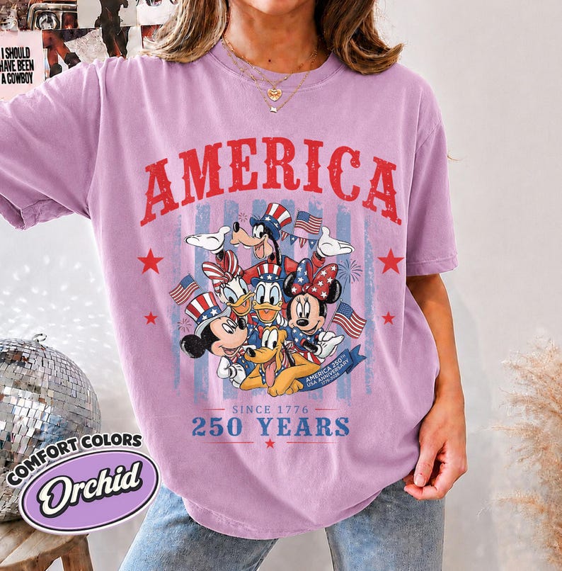 May include: An orchid-colored short-sleeve t-shirt featuring a graphic print of cartoon characters wearing patriotic hats. The text reads AMERICA, SINCE 1776, 250 YEARS, and AMERICA 250TH USA ANNIVERSARY. The design includes American flags and red stars.
