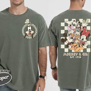 May include: Olive green t-shirt featuring Goofy on the front and a group of Disney characters on the back. The back design includes Mickey Mouse, Minnie Mouse, Donald Duck, Daisy Duck, Pluto, and Goofy. The text "Mickey & Co. EST. 1928" is also visible.