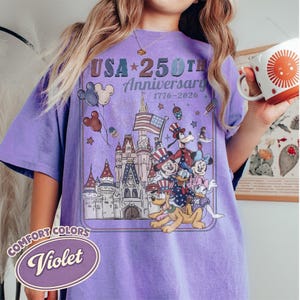 Puede incluir: Camiseta violeta Comfort Colors con un dise&ntilde;o gr&aacute;fico que celebra el 250 aniversario de EE. UU. El dise&ntilde;o incluye un castillo, fuegos artificiales, la bandera estadounidense y personajes de Disney.