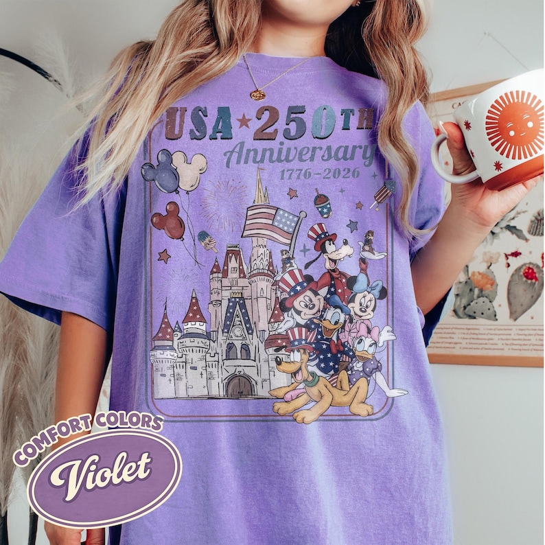 Puede incluir: Camiseta violeta Comfort Colors con un dise&ntilde;o gr&aacute;fico que celebra el 250 aniversario de EE. UU. El dise&ntilde;o incluye un castillo, fuegos artificiales, la bandera estadounidense y personajes de Disney.