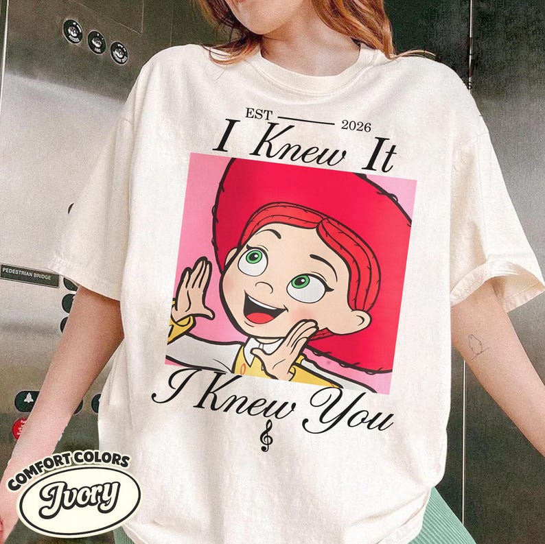 May include: An ivory short-sleeve t-shirt featuring a graphic of a red-haired character in a red hat. The text above reads EST 2026 I Knew It and the text below reads I Knew You with a musical note symbol. The shirt has a crew neckline and loose fit.