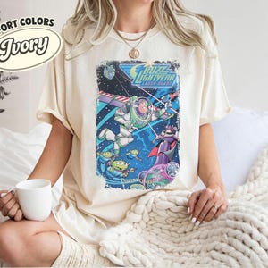 Pode incluir: Uma t-shirt cor de marfim com uma impress&atilde;o gr&aacute;fica de Buzz Lightyear Laser Blast, apresentando Buzz Lightyear, Zurg e alien&iacute;genas verdes no espa&ccedil;o. O texto diz Buzz Lightyear Laser Blast e Disneyland. Etiqueta Comfort Colors Ivory vis&iacute;vel.