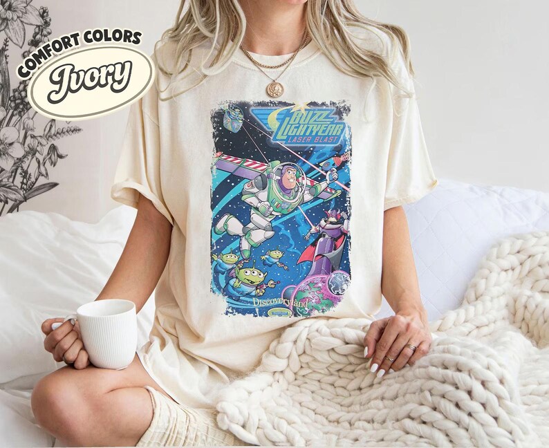 Pode incluir: Uma t-shirt cor de marfim com uma impress&atilde;o gr&aacute;fica de Buzz Lightyear Laser Blast, apresentando Buzz Lightyear, Zurg e alien&iacute;genas verdes no espa&ccedil;o. O texto diz Buzz Lightyear Laser Blast e Disneyland. Etiqueta Comfort Colors Ivory vis&iacute;vel.