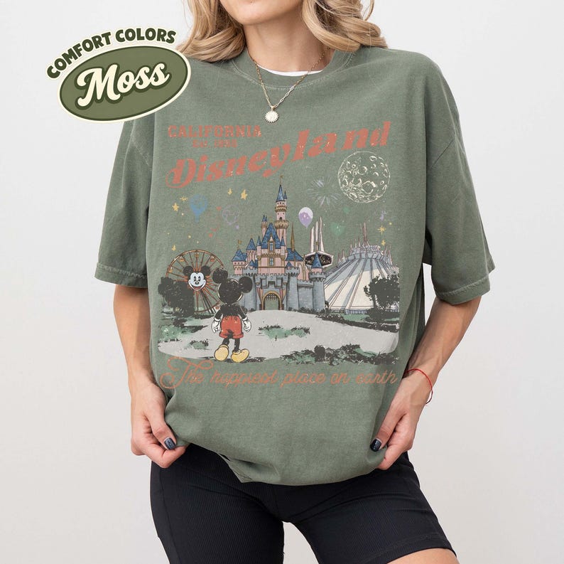 May include: Moss green t-shirt with a Disneyland graphic. The design features the Disneyland castle, Mickey Mouse, and the text "California Disneyland" and "The happiest place on earth". The shirt has a vintage look.