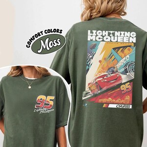 May include: Moss green t-shirt featuring Lightning McQueen from the Cars movie. The front displays "95" and "Lightning McQueen" text. The back showcases a racing scene with McQueen, the number "95", and the text "Lightning McQueen Racing Series".