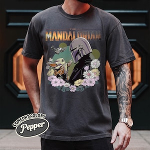 May include: Dark gray t-shirt featuring a graphic of The Mandalorian and Grogu surrounded by flowers. The text "THE MANDALORIAN" is above the image. The shirt has a Comfort Colors Pepper label.