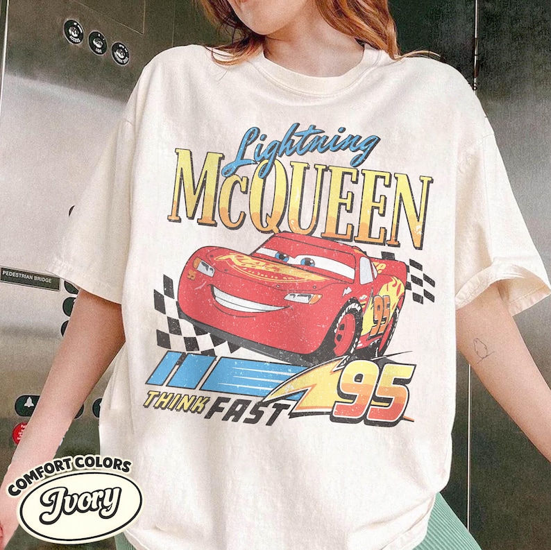 K&ouml;nnte beinhalten: Elfenbeinfarbenes T-Shirt mit einem Aufdruck von Lightning McQueen aus dem Film Cars. Das Design enth&auml;lt den Text "Lightning McQueen", "Think Fast" und die Nummer "95". Das Shirt ist von der Marke Comfort Colors.