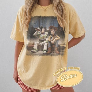 May include: Yellow Comfort Colors t-shirt featuring a distressed graphic of Buzz Lightyear and Woody from Toy Story. The graphic shows the characters sitting together. The shirt has a relaxed fit and the words "Comfort Colors Butter" are printed on the bottom right.