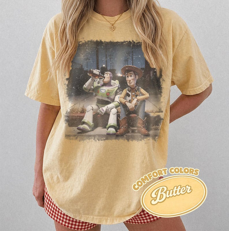 May include: Yellow Comfort Colors t-shirt featuring a distressed graphic of Buzz Lightyear and Woody from Toy Story. The graphic shows the characters sitting together. The shirt has a relaxed fit and the words "Comfort Colors Butter" are printed on the bottom right.