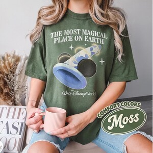K&ouml;nnte beinhalten: Olivgr&uuml;nes T-Shirt mit dem Text "THE MOST MAGICAL PLACE ON EARTH" und einer Grafik eines Zauberhuts und Mickey Mouse Ohren. Das Shirt hat auch die Worte "Walt Disney World" unter der Grafik aufgedruckt. Das Shirt ist mit "Comfort Colors" und "Moss" gekennzeichnet.