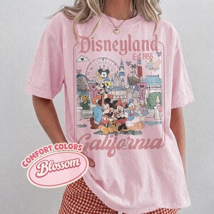 Może przedstawiać: R&oacute;żowa koszulka z grafiką Disneyland. Grafika przedstawia Myszka Miki, Minnie, Kaczora Donalda i inne postacie Disneya przed diabelskim młynem i zamkiem. Widoczny jest r&oacute;wnież napis "Disneyland California Est. 1955".