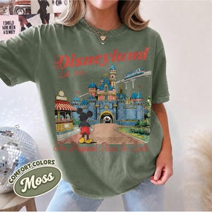 May include: A moss green t-shirt featuring a graphic print of a blue castle, a monorail, and Mickey Mouse. The text reads Disneyland Est. 1955 and The Happiest Place On Earth in red lettering. The shirt has short sleeves and a crew neckline.
