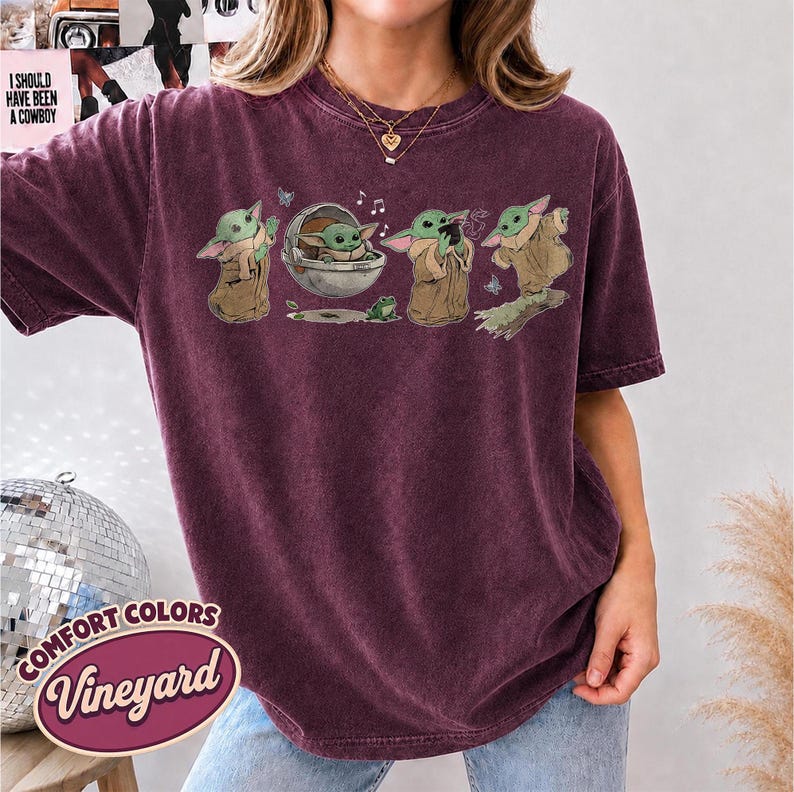 May include: A dark maroon short-sleeve t-shirt featuring a graphic print of a small green creature in various poses, including sitting in a floating pod, holding a cup, and standing, accompanied by a small green frog and musical notes.