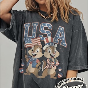 May include: Dark gray oversized t-shirt featuring the word "USA" in blue and red, with stars. Two cartoon chipmunks are dressed in patriotic attire, one holding an American flag. The shirt has the text "COMFORT COLORS Pepper".