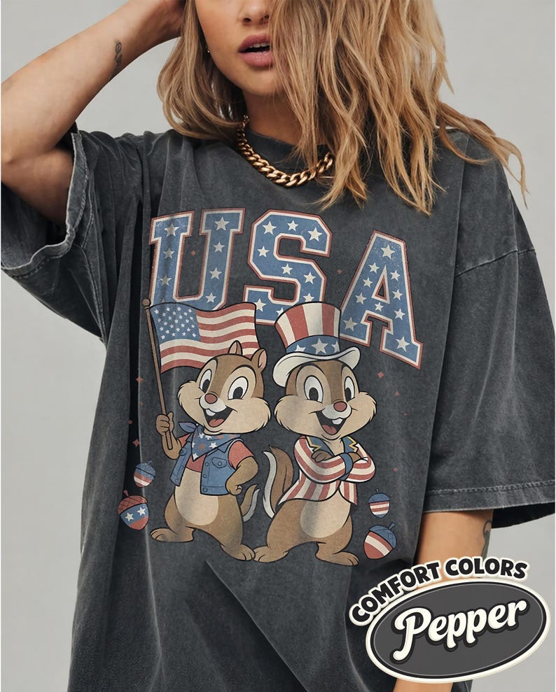 May include: Dark gray oversized t-shirt featuring the word "USA" in blue and red, with stars. Two cartoon chipmunks are dressed in patriotic attire, one holding an American flag. The shirt has the text "COMFORT COLORS Pepper".