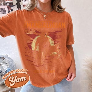 May include: A burnt orange short-sleeve t-shirt featuring a distressed graphic print of The Mandalorian and Grogu against a sunset background. The text THE MANDALORIAN is printed above the figures, and THIS IS THE WAY is printed below in a vintage style.