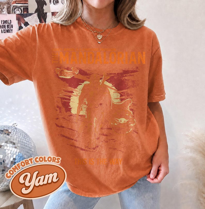 May include: A burnt orange short-sleeve t-shirt featuring a distressed graphic print of The Mandalorian and Grogu against a sunset background. The text THE MANDALORIAN is printed above the figures, and THIS IS THE WAY is printed below in a vintage style.
