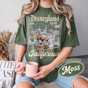 Może przedstawiać: Zielona koszulka z grafiką Disneyland z Myszką Miki, Minnie, Kaczorem Donaldem i innymi postaciami. Na koszulce nadrukowano napis "Disneyland California Est. 1955". Koszulka jest w kolorze Comfort Colors Moss.