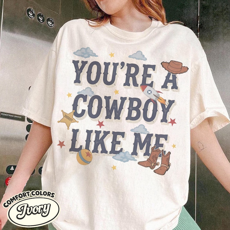 May include: An ivory short-sleeve t-shirt featuring the text "YOU'RE A COWBOY LIKE ME" in dark blue letters. The design includes illustrations of a brown cowboy hat, a rocket, a yellow sheriff star, a colorful ball, and brown cowboy boots.