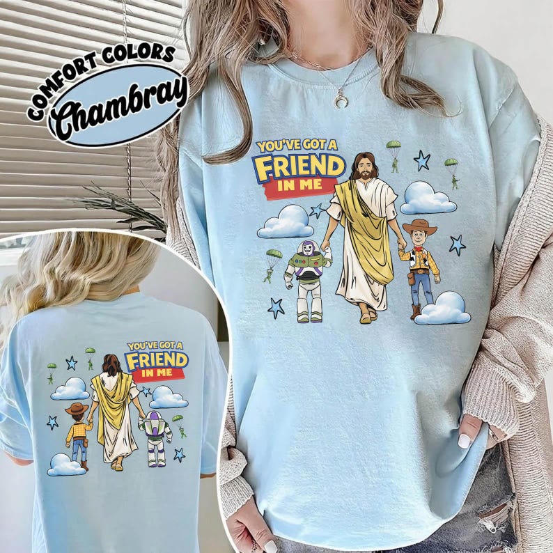 May include: Pale blue t-shirt featuring the text "YOU'VE GOT A FRIEND IN ME" and a graphic of Jesus with Buzz Lightyear and Woody. The design includes clouds, stars, and small parachutes.
