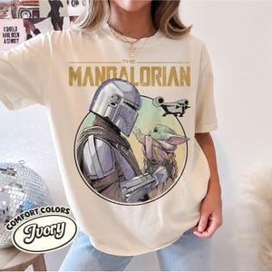 Puede incluir: Una camiseta de manga corta color marfil con un estampado gr&aacute;fico de The Mandalorian y Grogu. El dise&ntilde;o incluye el texto THE MANDALORIAN en dorado, un marco circular y la silueta de una nave espacial. La camiseta tiene cuello redondo y corte holgado.