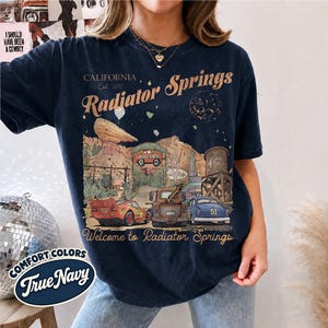 May include: A dark navy blue short-sleeve t-shirt featuring a graphic print of Radiator Springs with text that reads California Est 2012 Radiator Springs Welcome to Radiator Springs, depicting a red race car, a tow truck, and a blue car.