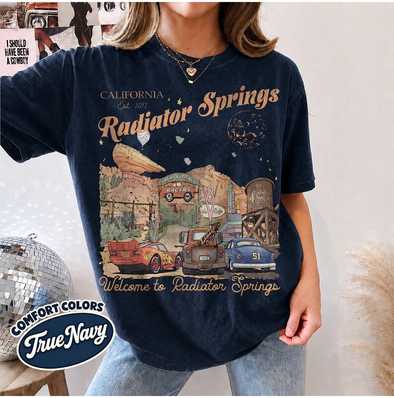 May include: A dark navy blue short-sleeve t-shirt featuring a graphic print of Radiator Springs with text that reads California Est 2012 Radiator Springs Welcome to Radiator Springs, depicting a red race car, a tow truck, and a blue car.