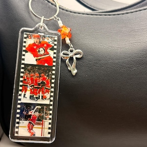 May include: A clear acrylic keychain featuring hockey game images. The keychain has a silver ring, orange star-shaped bead, white beads, and a silver bow charm. The background is a black purse.