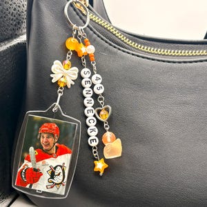 May include: A clear acrylic keychain featuring a hockey player in a red helmet and jersey. The keychain is adorned with orange and white beads, a bow, and the name "SENNECKE" spelled out with letter beads. A black purse is in the background.