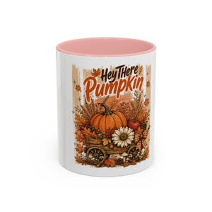 May include: A white ceramic mug with a soft pink interior rim featuring a graphic illustration of a wooden wagon filled with a large orange pumpkin, a white daisy, autumn leaves, and wheat. The text Hey There Pumpkin is printed above.