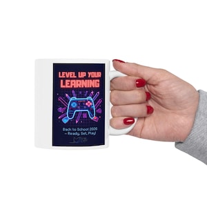 May include: A white ceramic mug featuring a dark blue rectangular graphic with neon-style illustrations of a game controller and school supplies. The text reads: LEVEL UP YOUR LEARNING, Back to School 2026 – Ready, Set, Play! in bright pink and blue.