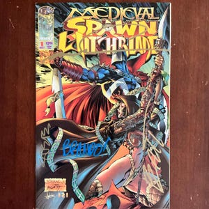 May include: A comic book titled Medieval Spawn Witchblade issue number 1, featuring a colourful illustration of two armoured characters in a battle scene. The cover includes bold yellow text, multiple signatures in blue and gold ink, and an Image logo.