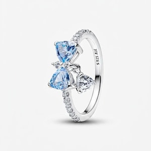 May include: A silver ring featuring a bow design with two light blue heart-shaped gemstones and a dangling clear heart-shaped gemstone. The ring band is encrusted with small clear gemstones. The ring is stamped with "S 925 ALE".