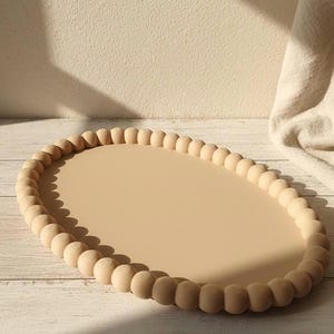 May include: An oval decorative tray with a light beige surface and a border of wooden beads. The tray is set on a light-coloured wooden surface, with a neutral background. The tray's dimensions are approximately 38 cm long and 25 cm wide.