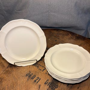May include: A set of white ceramic plates with a scalloped edge design. One plate is displayed on a metal stand, while a stack of plates rests on a wooden surface. The plates have a classic, elegant look.